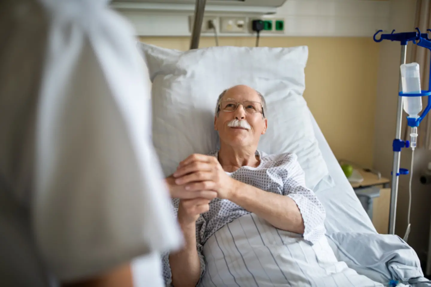 Ein älterer Mann liegt in einem Krankenhausbett und hält die Hand eines Pflegers